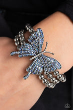 Load image into Gallery viewer, Strung along elastic stretchy bands, a trio of silver and textured silver beads and accents wrap around the wrist. Featured atop the beaded collection, an oversized silver butterfly, with intricate details, is sprinkled with dainty blue iridescent rhinestones across its wings and body, for a dramatically dazzling finish. Due to its prismatic palette, color may vary. Sold as one individual bracelet.