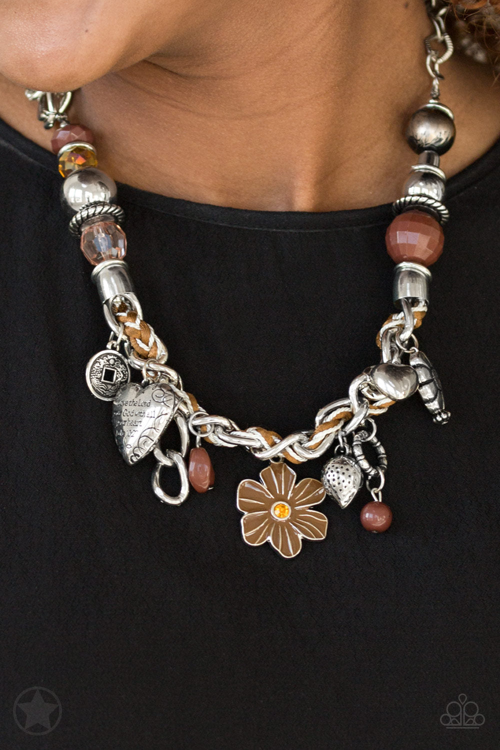 Brown and ivory cording is braided through a chunky silver chain. A unique variety of charms decorate the Blockbuster piece, including a delicate flower and a heart inscribed with the phrase 