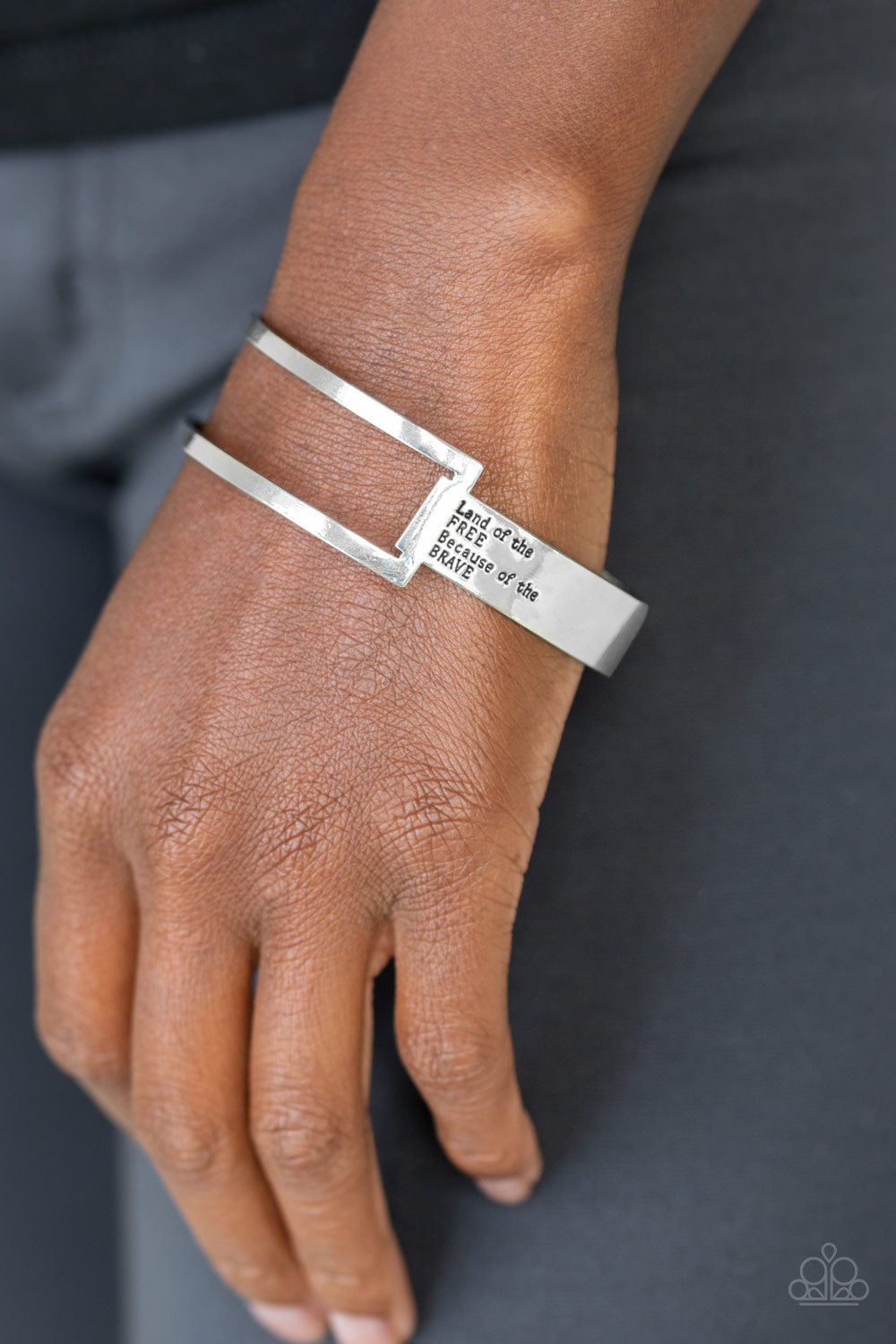 An airy rectangular frame attaches to a flat silver bar that curves around the wrist and is stamped in the phrase, “Land of the free because of the brave,” creating a patriotic inspired cuff. Sold as one individual.