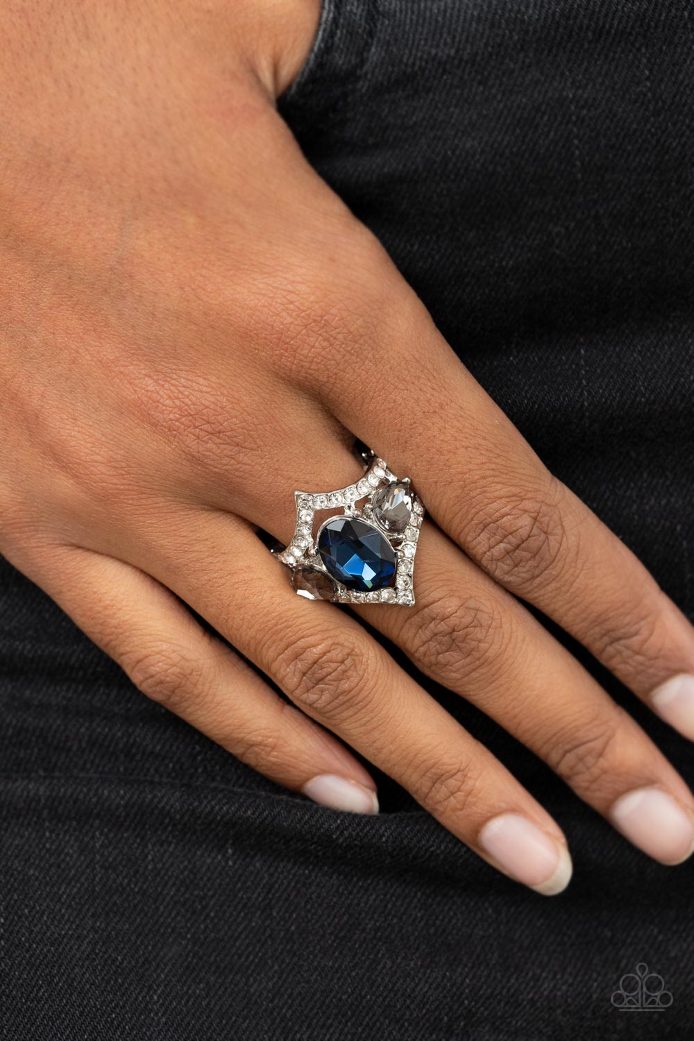 An oversized blue oval gem is flanked by a pair of smoky teardrop rhinestones atop a white rhinestone dotted frame, resulting in a dramatic dazzle atop the finger. Features a stretchy band for a flexible fit.  Sold as one individual ring.