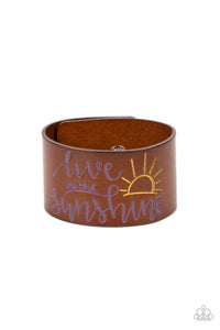 A thick band of light brown leather is stamped with the cheerful phrase, "live in the sunshine," along with a radiating sun design. The purple font complements the yellow tint of the sun in an optimistic finish. Features an adjustable snap closure.  Sold as one individual bracelet.