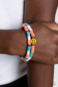 Coral, blue, and white rubber discs combine with sporadically placed silver beads to provide a vibrant pop of color along the wrist. Meeting in the center of one of the multi-shaded bracelets, a yellow bead with a smiley face, featuring stars for eyes, rests for a youthful finish.  Sold as one set of two bracelets.