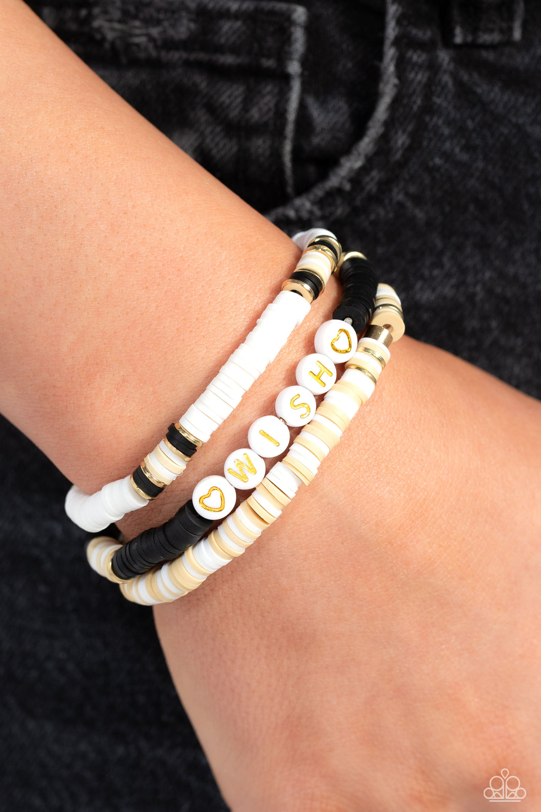 Varying in shades, tan, black, and white clay discs pair with shiny gold disc beads and accents creating layers across the wrist. Featured on one of the bracelets, white beads stamped with gold lettering spell out the word 
