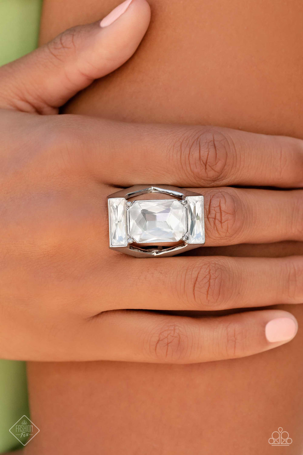An exaggerated, oversized, rectangular frame encases a large, white, emerald-cut gem, showcasing its scintillating shimmer. Skinny white gems chiseled into rectangular bars are pressed into the top and bottom of the industrial frame, resulting in an unapologetic statement piece with unmatched sparkle. Features a stretchy band for a flexible fit.  Sold as one individual ring.