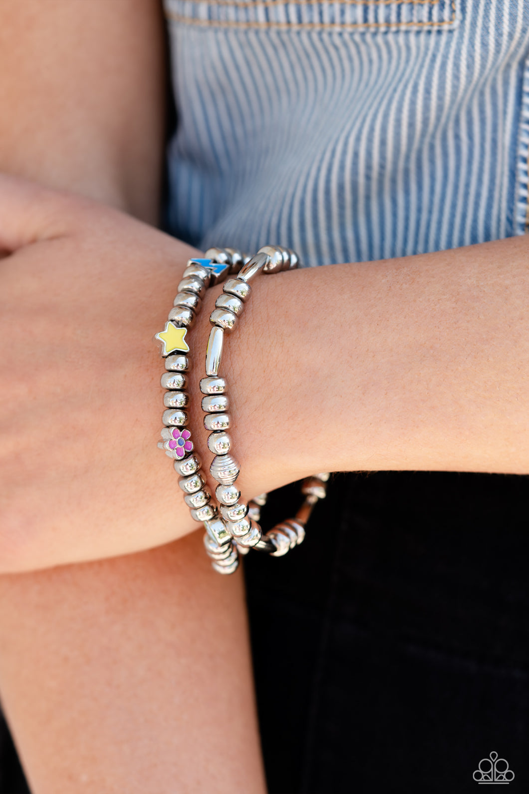 A strand of textured, cylindrical, and round high-sheen silver beads combine with a single strand of silver beads, sporadically infused with vivacious charm beads to create an energetic stack of stretchy bracelets. The colorful beaded stack features a yellow star, blue lightning bolt, green money bag, orange tropical flower, red lips, blue and green earth, pink and purple flower, and a pink heart charm with the words 
