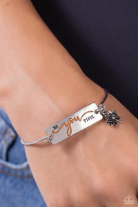 The word "BeYOUtiful" is stamped across a thick plate of silver, with the "YOU" in a playful orange font. A textured, silver lotus flower charm gathers at the side of the inspirational centerpiece as it connects to a skinny silver bar that wraps around the wrist. Features a hook and eye closure.  Sold as one individual bracelet.
